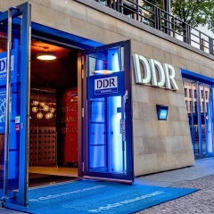 Entrance area DDR Museum. The doors are wide open, illuminated with blue lights on the right and left. Blue carpet lies on the floor. Large white GDR letters hang on the wall.