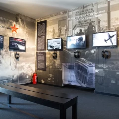 View of the exhibition space in The Wall Museum. Televisions with headphones hang on the wall, a bench stands in the room and text panels hang on the wall.