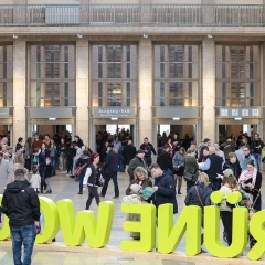 View into the rooms of Messe Berlin. In the foreground are green letters that spell out “Internationale Grüne Woche Berlin”. Many visitors walk from right to left. In the background are five doors through which visitors stream.