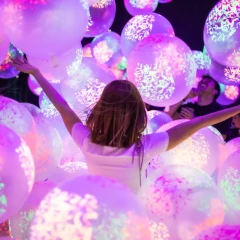 View of the exhibition room in the Balloon Museum. Lots of different pink balloons that light up. In the middle of it all, a girl stands with her back to the camera and stretches her arms up in the air.