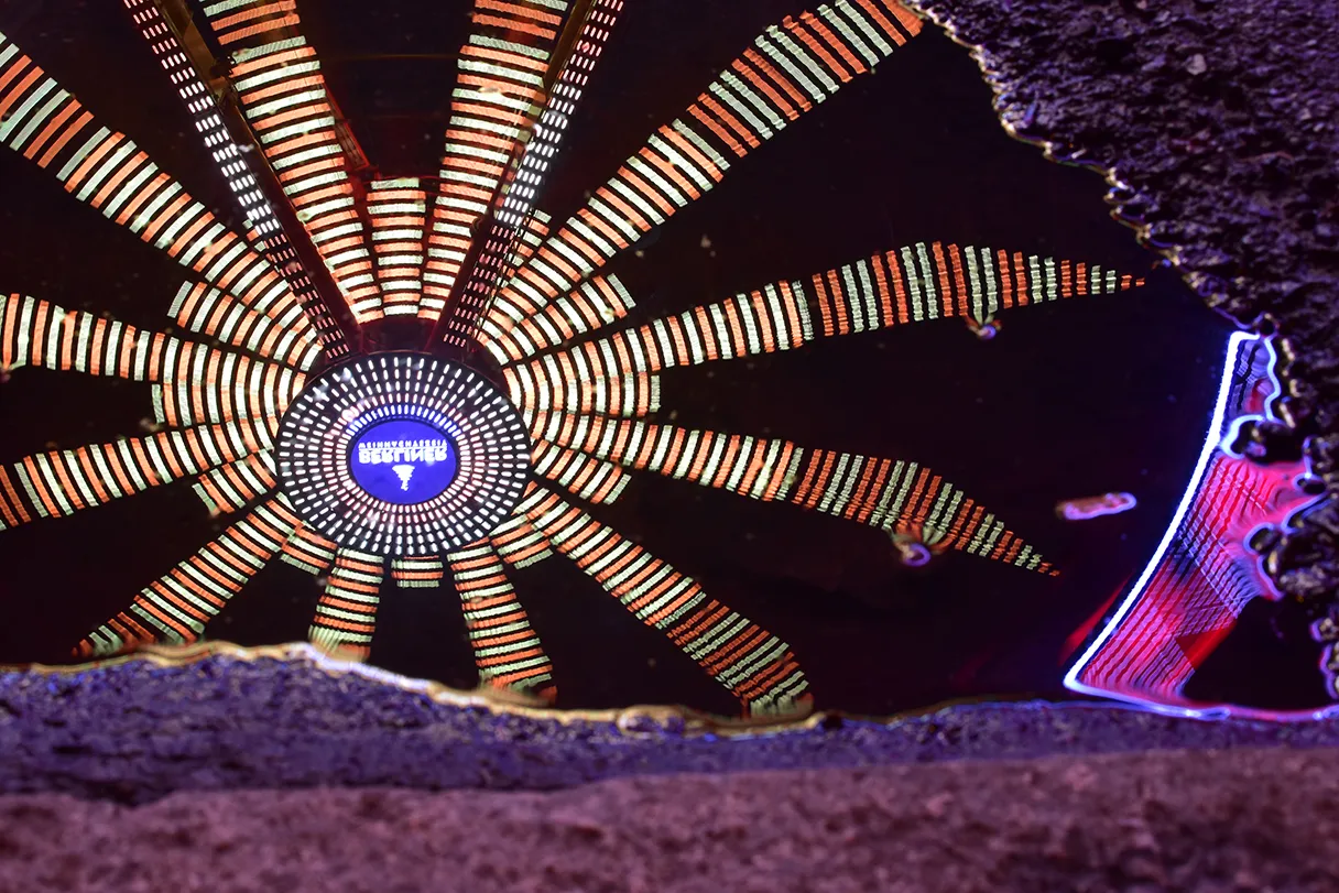 The Ferris wheel from the Christmas market is reflected in a puddle. It is dark and the lights are shining in the water.