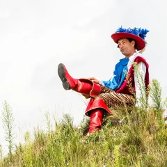 A man in a costume is sitting on a green hill. It is supposed to represent Puss in Boots as a human. The man is wearing large red leather boots, a red hat with blue feathers and a red vest.