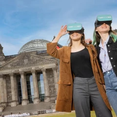 Two women are wearing VR goggles and standing on the lawn in front of the Reichstag in Berlin.