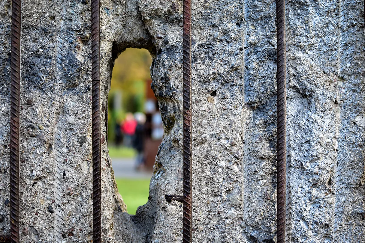 View through a hole in the Berlin Wall. In the background, people, grass and green trees can be seen out of focus. The iron struts that were anchored in the concrete wall can be seen in the foreground.