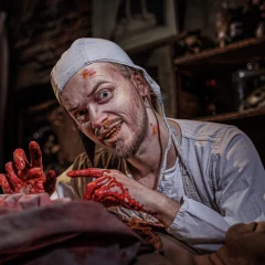 Berlin Dungeon, man with bloodstained hands stands behind a corpse and looks confused into the camera, he wears a white coat, cap and face mask around his neck