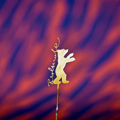 The golden bear of the Berlinale is pinned to a stick on which Berlinale is written in curved lettering. The background is patterned red and blue.