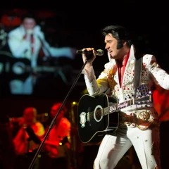 An Elvis Presly lookalike stands on stage in the typical white Elvis suit. He has a black guitar slung around his neck and holds the microphone firmly in his hand while he sings.