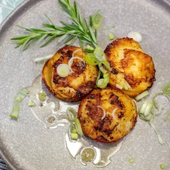 Essensteller aus der Vogelperspektive. Es sind drei Kartoffelpuffer mit etwas Lauchzwiebeln und einem Rosmarinzweig abgebildet. Oben links ist noch etwas silbernes Besteck zu erkennen.