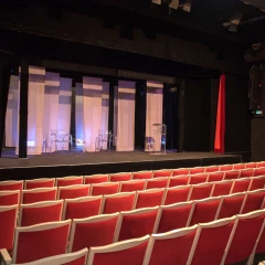 View into the auditorium of the Berlin Crime Theater. Red chairs are lined up next to each other. Various white curtains and glass cabinets have been placed on the stage. The auditorium is empty, no people to be seen.