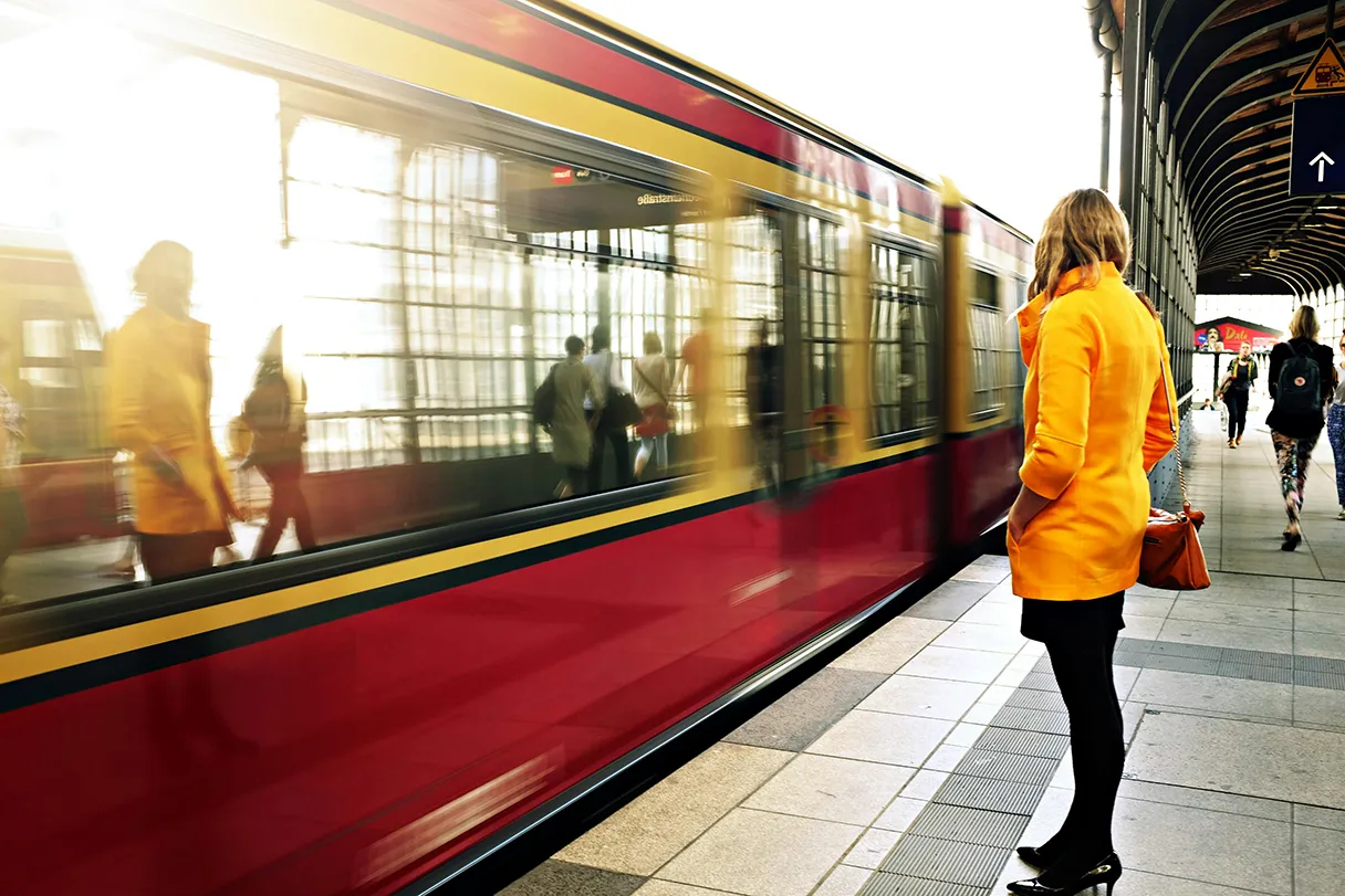 A woman in a yellow coat, black skirt and shoes is standing at the station. She looks towards the arriving suburban train, which looks somewhat blurred by the movement. The windows reflect the windows of the station.