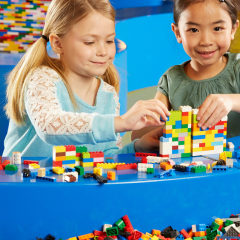 Lego Discovery Centre Zwei Mädchen spielen mit bunten Legosteinen und haben Spaß. Ein Mädchen guckt in die Kamera und lacht und das andere Mädchen steckt konzentriert die Steine zusammen.