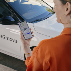 Eine Frau schaut auf ihr Handy und die App von free2move. Im Hintergrund ist ein weißes free2move Auto zu sehen.