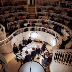 Im Tieranatomischen Theater sitzen Zuschauer:innen und schauen nach unten, um zu sehen, was dort am Tisch passiert. Dort sitzen 8 Personen an einem Tisch und unterhalten sich.