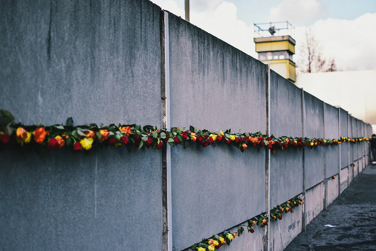 View of the Berlin Wall at the memorial in Bernauer Strasse. The Wall is decorated with red, yellow and orange roses. In the distance, you can see a border tower peeking over the Wall on the right edge of the picture.