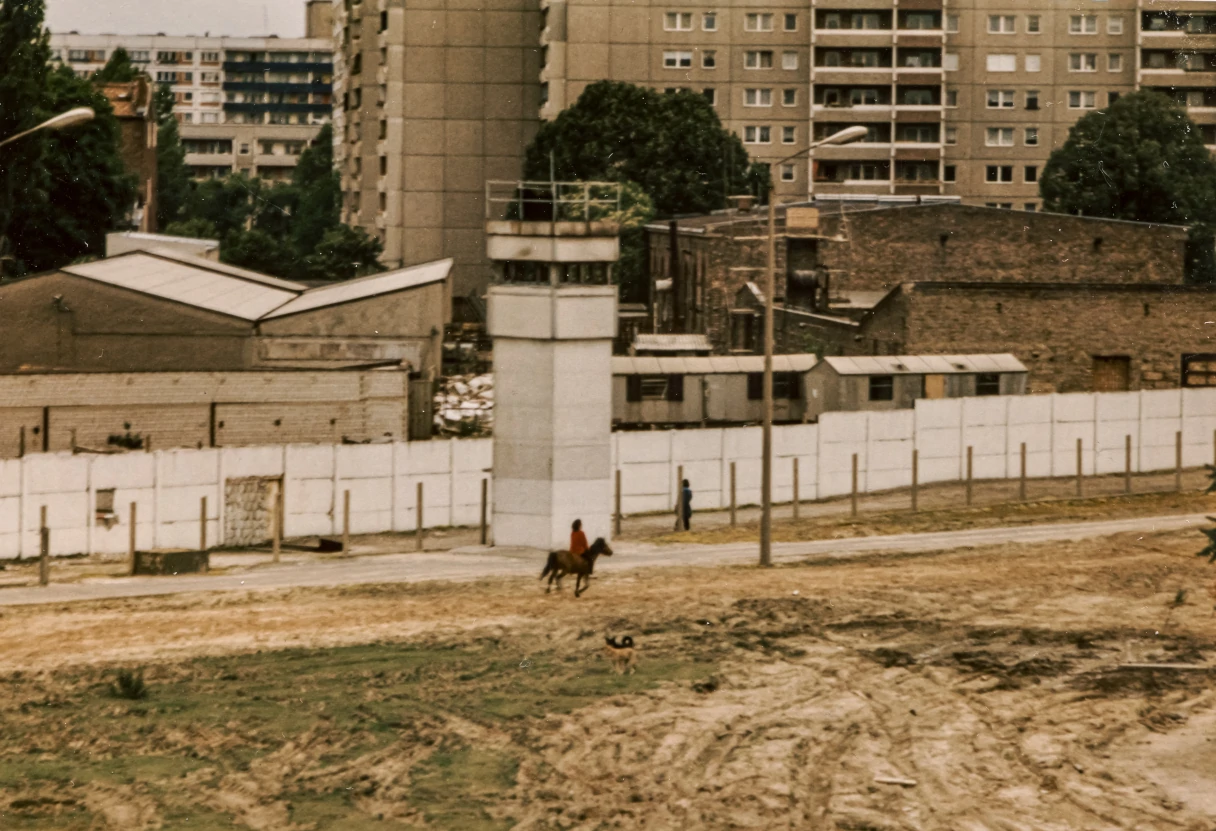 Analog picture. View of the border strip from a residential building. Lots of sand, wall, border tower, prefabricated building in the background. A person is riding a horse along the Wall.