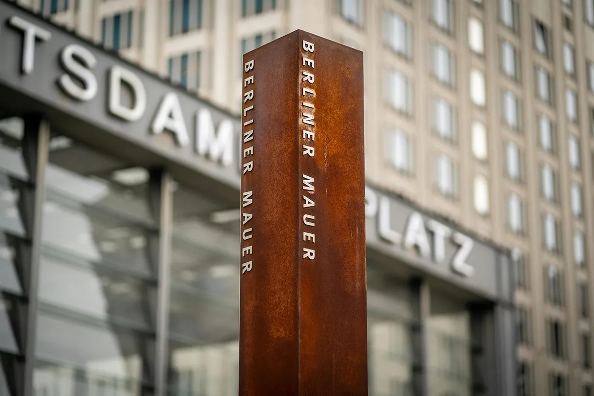 Rust-brown stele with the Berlin Wall written on it is placed in the center of the picture. Potsdamer Platz station and a skyscraper can be seen in the background.