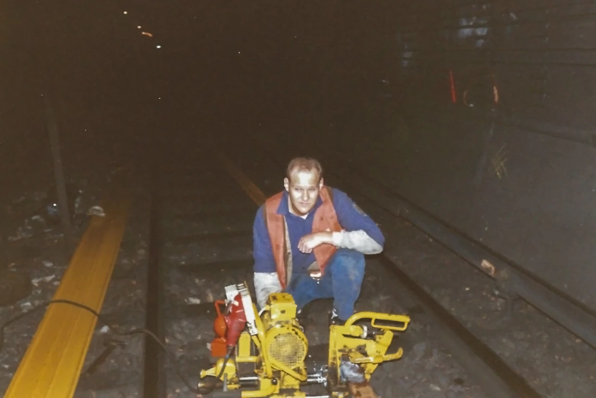 Analog image. Matthias in overalls at work in a dark subway tunnel. He is crouching between the tracks and there are yellow devices in front of him.