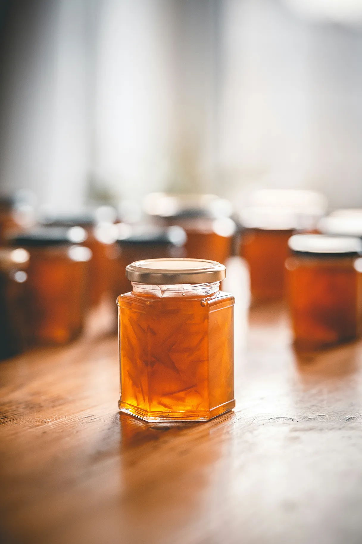 Many jars of orange marmalade stand on a table. One glass is in the foreground and is the focus. Warm tones. You can still see the orange slices in the glass.