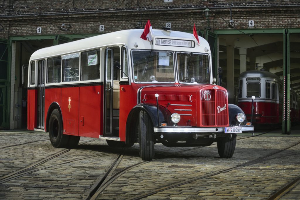 An old Wiener Linien bus is parked in the depot. Old streetcars and tracks can be seen in the background.