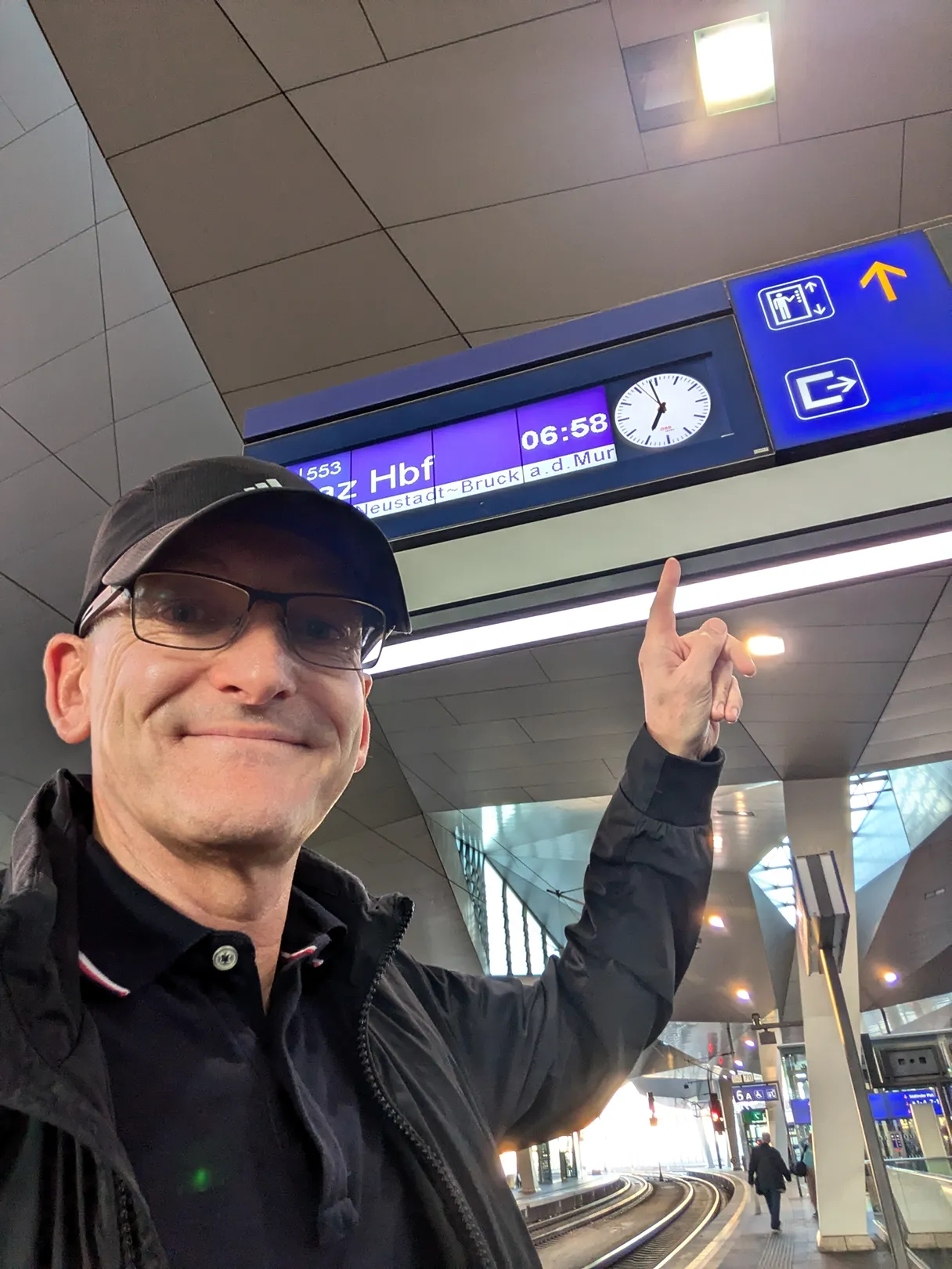 Angekommen in Wien, Olaf steht auf dem Bahnsteig und zeigt mit dem Finger auf die Uhr, es ist sieben Uhr morgens, er lächelt in die Kamera