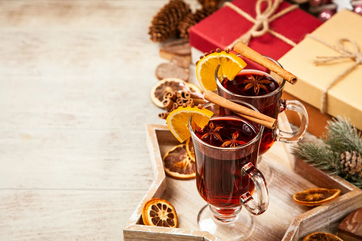 Two glasses of mulled wine are placed on a wooden star tray. Orange slices are hung on the glasses as Christmas decorations. Cinnamon sticks, dried orange slices and presents wrapped in red wrapping paper lie around them.