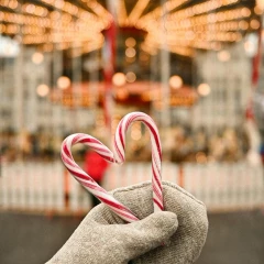 Eine Person hält mit einem Handschuh zwei rot-weiße Zuckerstangen in der Hand. Sie ergeben ein Herz. Im Hintergrund sieht man verschwommen ein altes Karussel auf dem Weihnachtsmarkt, welches hell erleuchtet ist.