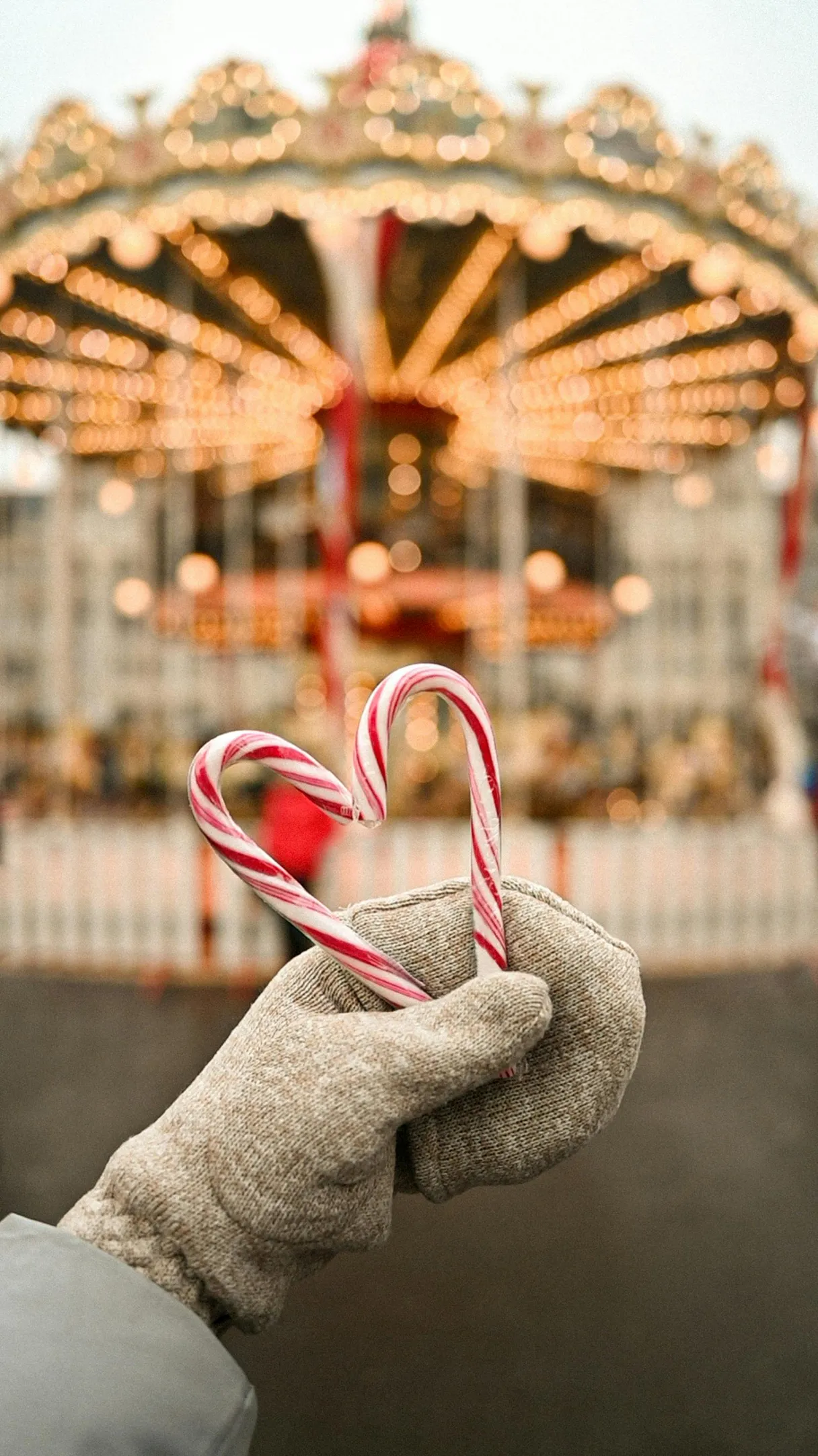 Eine Person hält mit einem Handschuh zwei rot-weiße Zuckerstangen in der Hand. Sie ergeben ein Herz. Im Hintergrund sieht man verschwommen ein altes Karussel auf dem Weihnachtsmarkt, welches hell erleuchtet ist.