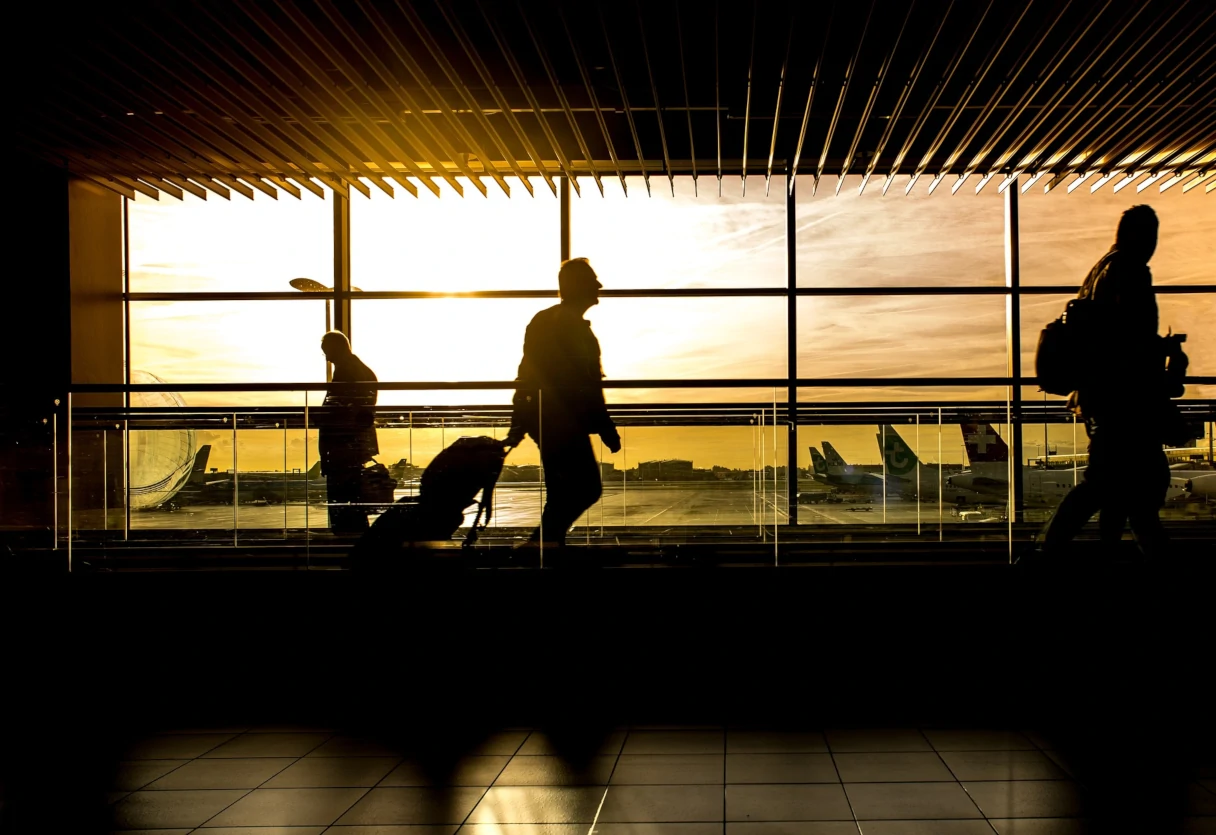 Airport, people walking across the conveyor belt with suitcases in their hands, silhouettes, sun shining directly in, airplanes in the background 