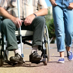 A man in a wheelchair holding a cane in his hand, next to him walks a woman in blue jeans and blue shoes.