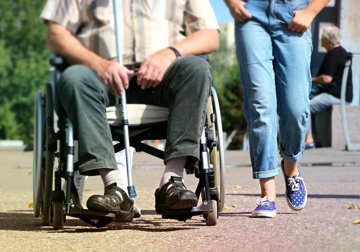 A man in a wheelchair holding a cane in his hand, next to him walks a woman in blue jeans and blue shoes.