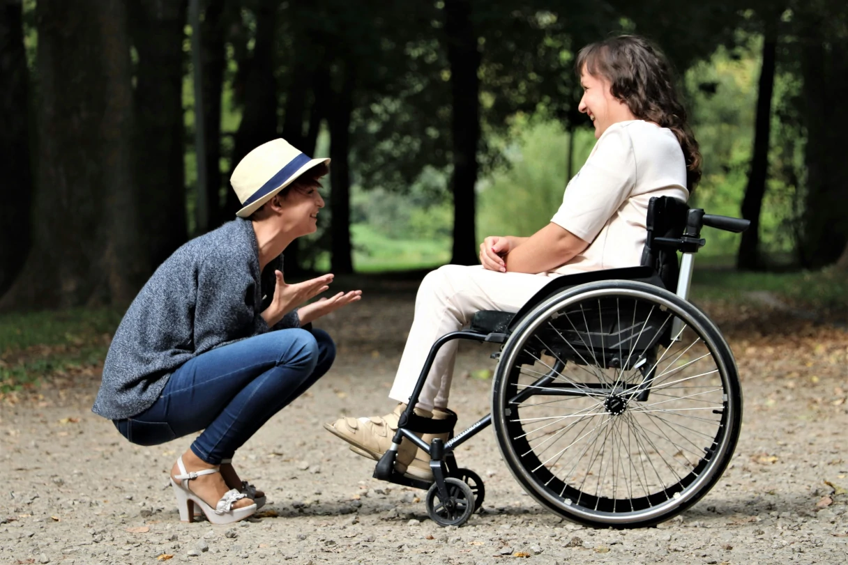 Woman sits in a wheelchair a woman with a hat squats in front of her, they both laugh