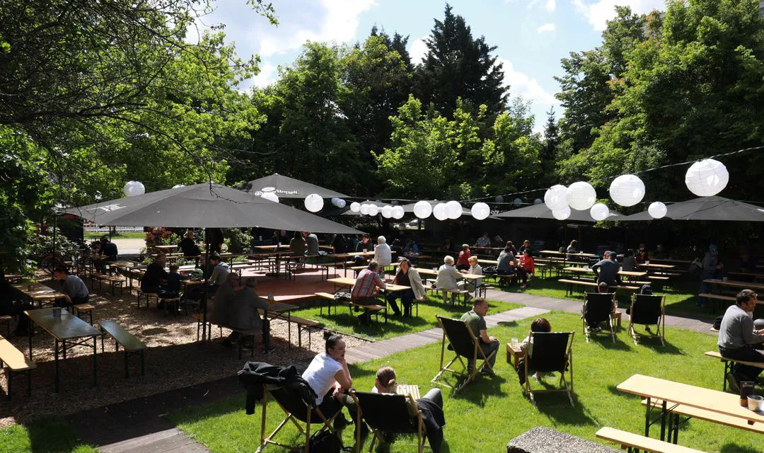 James June, Blick in den Biergarten, Tische und schware Sonnenschirme, weiße runde Lampions, grüner Rasen, Besucher sitzen in der Sonne in Liegestühlen