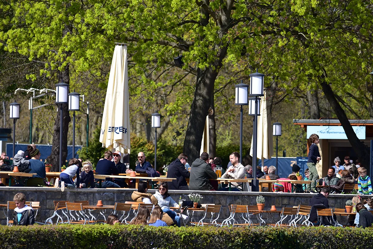 Zenner, Berlin, Blick vom Wasser aus auf den Biergarten, helle Schirme, die zusammengeklappt sind, Menschen sitzen an Tischen und unterhalten sich, grüner Baum im Hintergrund