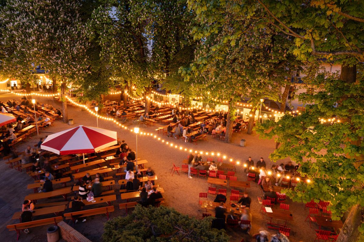 Blick von oben in den Garten des Biergarten Prater, Lichterketten hängen zwischen den Bäumen, Bierbänke mit Besuchern drauf, Nachtstimmung