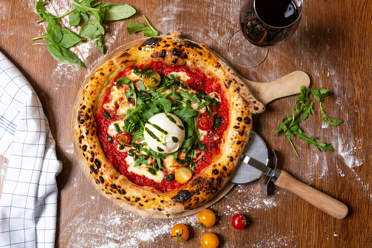 Pizza, Blick von oben auf die Burrata Pizza, mit Pizzaroller, Tomaten, Basilikum neben der Pizza