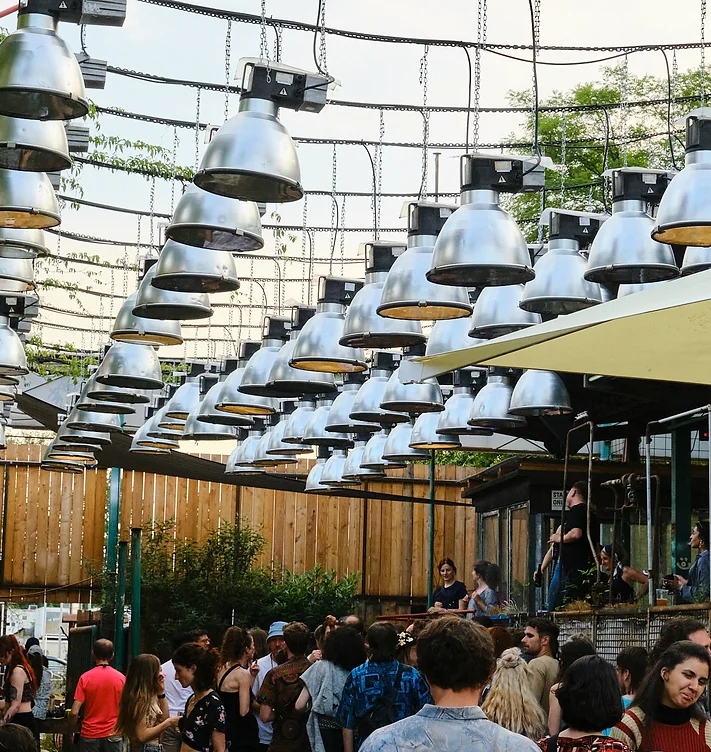 Biergarten, mehrere Reihen mit Metalllampen hängen im Biergarten, darunter sitzen viele Menschen