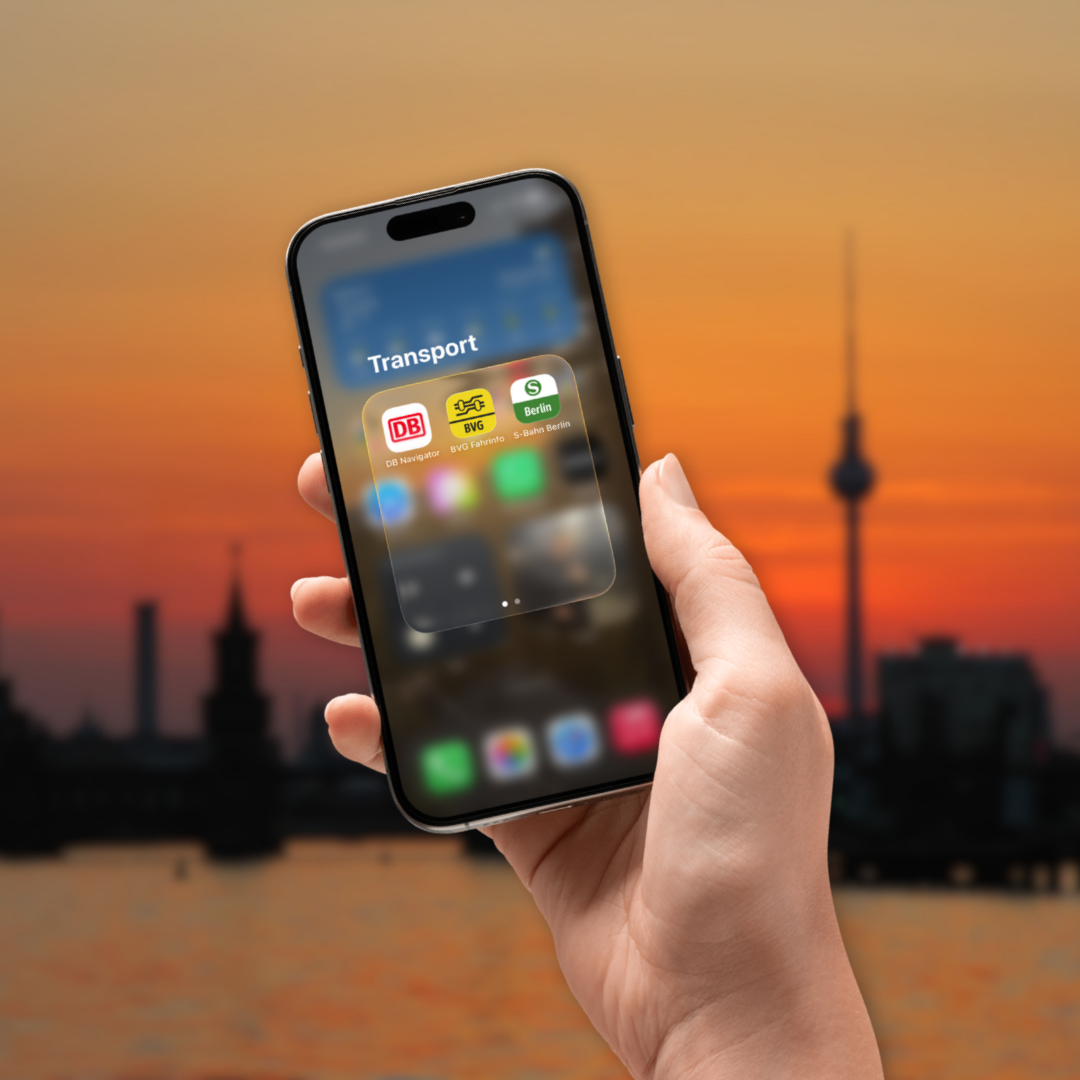 Silhouette von Berlin und der Oberbaumbrücke mit Fernsehturm im Sonnenuntergang. Im Vordergrund ist eine Hand zu sehen, die ein Handy hält. Es sind verschiedene Transport Apps zu sehen. Unter anderem der DB Navigator.