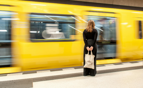 Eine Frau mit Jutebeutel steht vor einer einfahrenden gelben U-Bahn am U-Bahnhof.