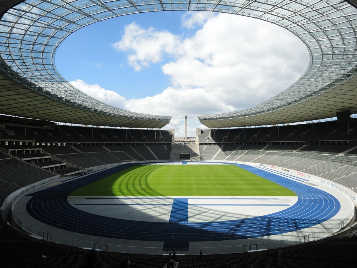 Blick in das Olympiastadion Berlin, rundes Dach, Blick in den blauen Himmel, blaue Tartanabahn, grüner Fußballrasen, ringsherum Sitzplätze