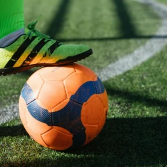 Soccer player stands with his yellow Adidas shoe on an orange soccer ball, which lies on the white line on the green soccer turf