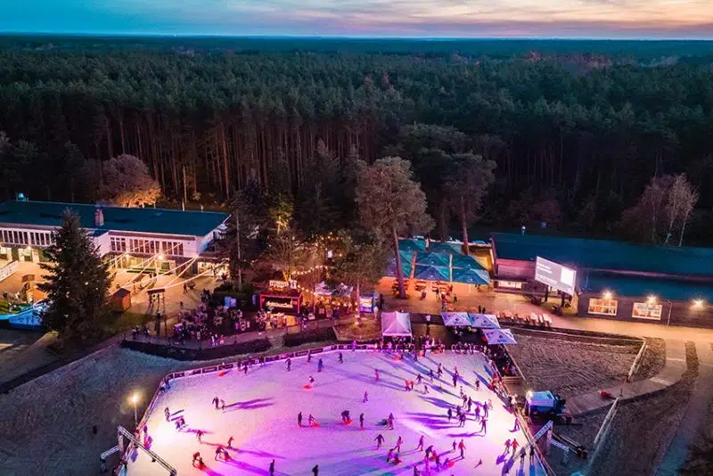 Vogelansicht auf die Winterwelt Grünau im Strandbad Grünau. Man sieht die große Eisbahn am Strand in pinkem Licht. Im Hintergrund ein großes Waldgebiet.