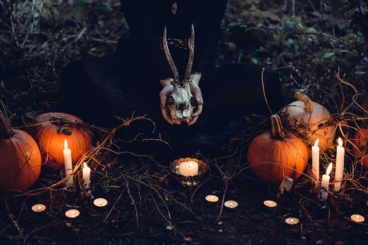 Halloween-Stimmung, eine Frau hält einen Tierschädel in den Händen, ringsherum sind Kürbisse, Kerzen aufgebaut, gruselige Stimmung, dunkles Bild