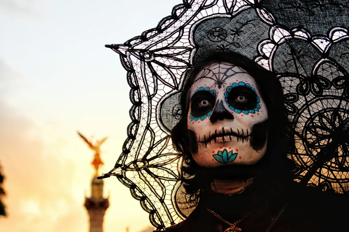 Halloween, scary made-up person with black lace umbrella and black clothes looks into the distance, in the background a high column with a figure as a silhouette can be recognized