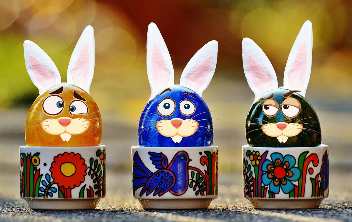 Three colorful Easter eggs, brown, blue and dark green, sit in colorful egg cups. The Easter eggs look like Easter bunnies as they have had faces and bunny ears glued on.