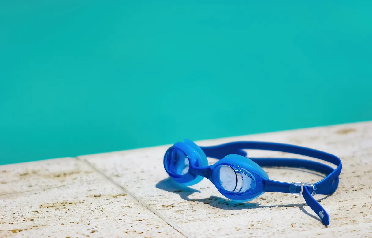 Schwimmbrille liegt am Beckenrad, grünes Wasser im Hintergrund