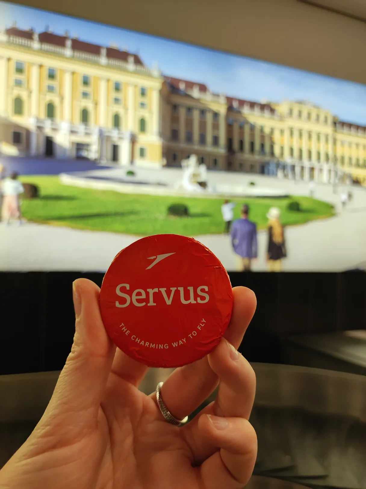 Schokoladentaler mit der Aufschrift "Servus" wird am Flughafen vorm Kofferband mit einer Hand hochgehalten, im Hintergrund ist das Schloss Schönrunn als Werbung zu sehen