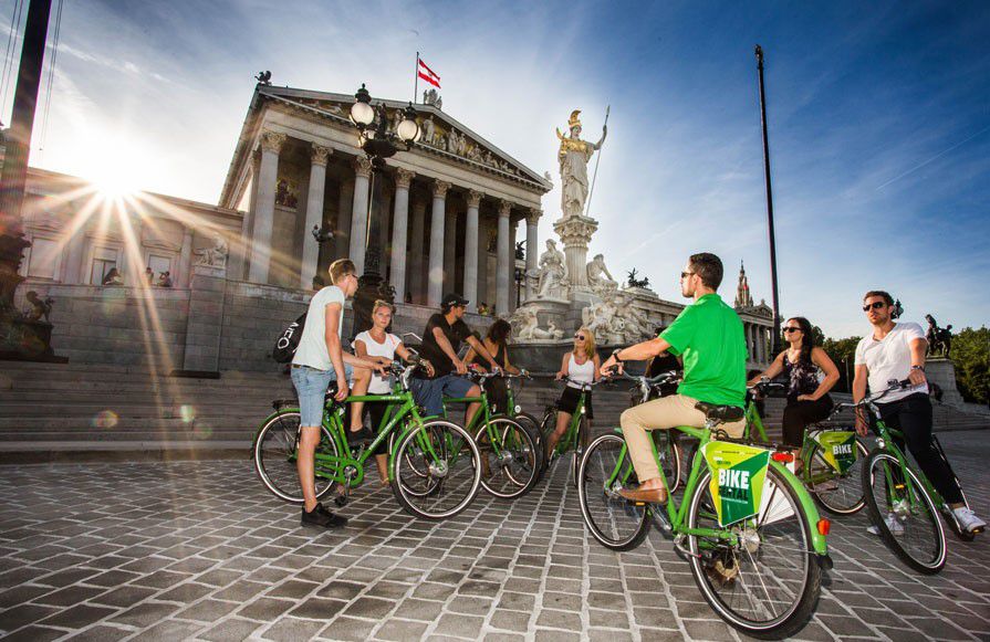 Fahrradtour mit Vienna Explorer Tours, Gruppe steht mit Fahrrädern  vorm Parlamentsgebäude, Sonnenstrahlen brechen links im Bild über die Gebäude