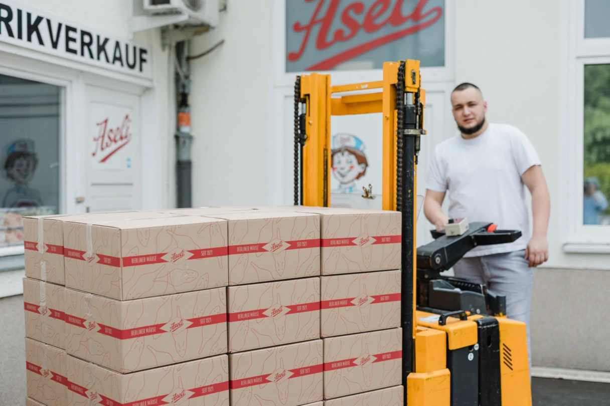 Aseli, Mann stitzt auf einem Gabelstapler auf dem beige Kisten mit dem Aseli Logo drauf gestapelt sind