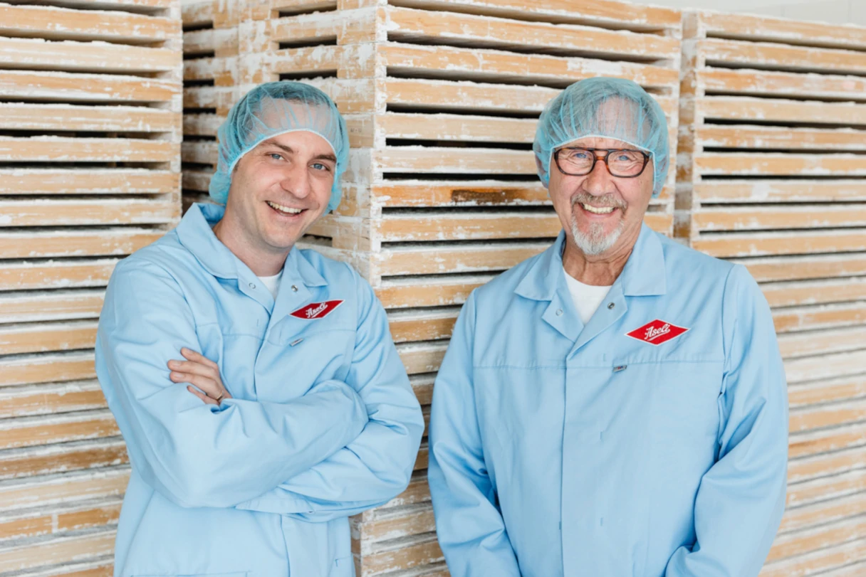 Aseli Manufaktur, Berlin, two men in blue coats and transparent hairnets on their heads stand in the factory, laughing