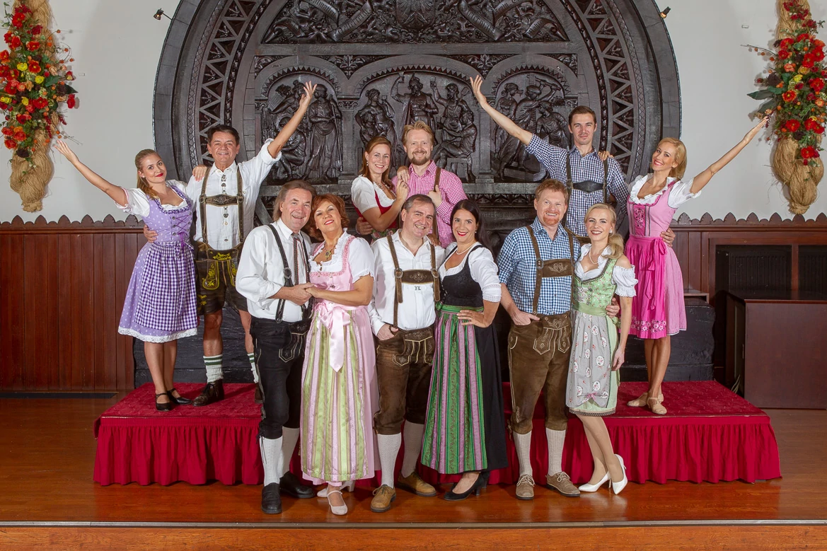 Austrian Dinner Show, Vienna, women in dirndls and men in lederhosen and shirts stand close together as a group, on a red pedestal, in front of a dark mural